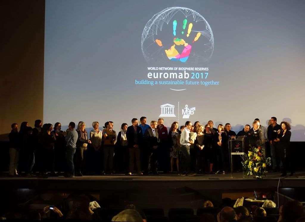 Kokouksen osanottajia ryhmäkuvassa esiintymislavalla. Taustalla heijastettuna Euromab -logo.